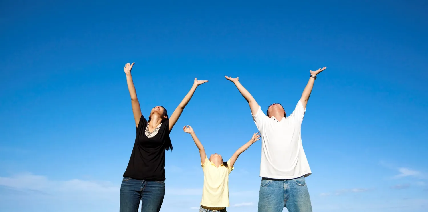 Family of three raising their arms together under a blue sky